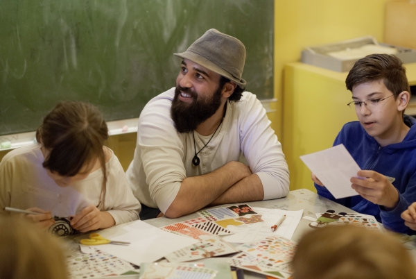 Ein junger Mann of Colour mit Hut sitzt an einem Tisch mit Kindern und bastelt.
