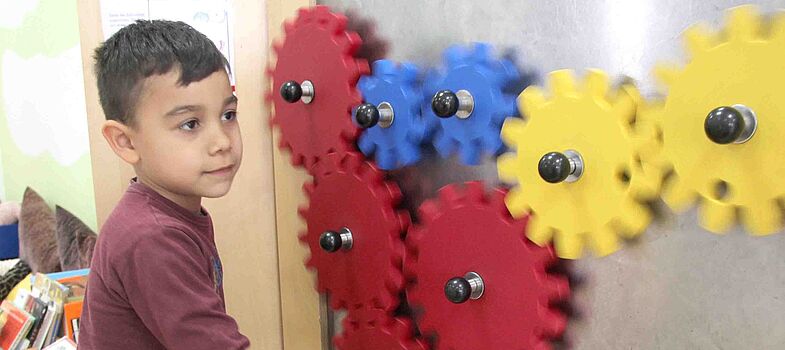 Ein Kind spielt an einer Wand, an der rote, blaue und gelbe Zahnräder, die ineinandergreifen, angebracht sind.